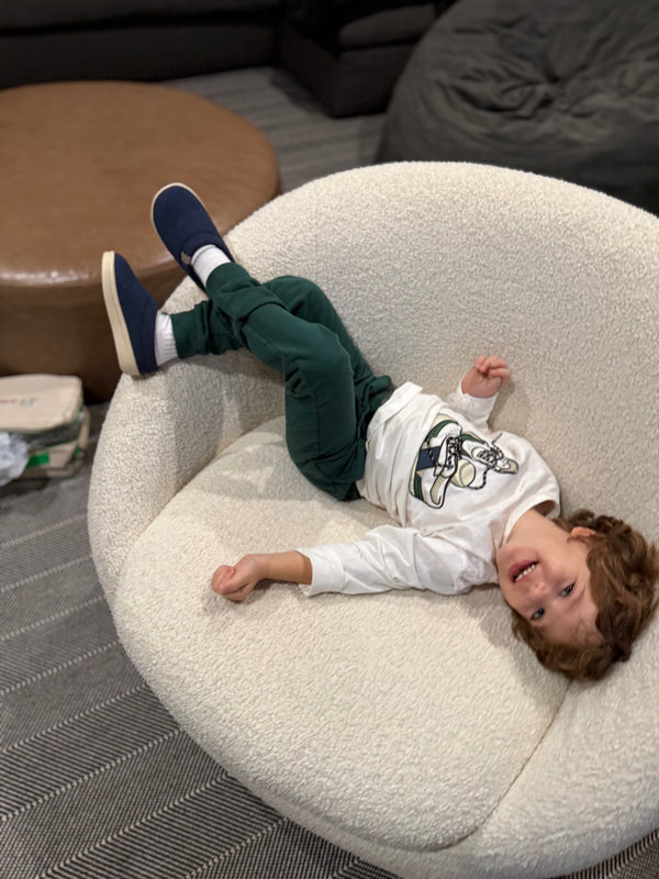 Angled view of Essential Kids slipper in navy on a child showcasing slip on upper and cream sole on a round cream boucle chair