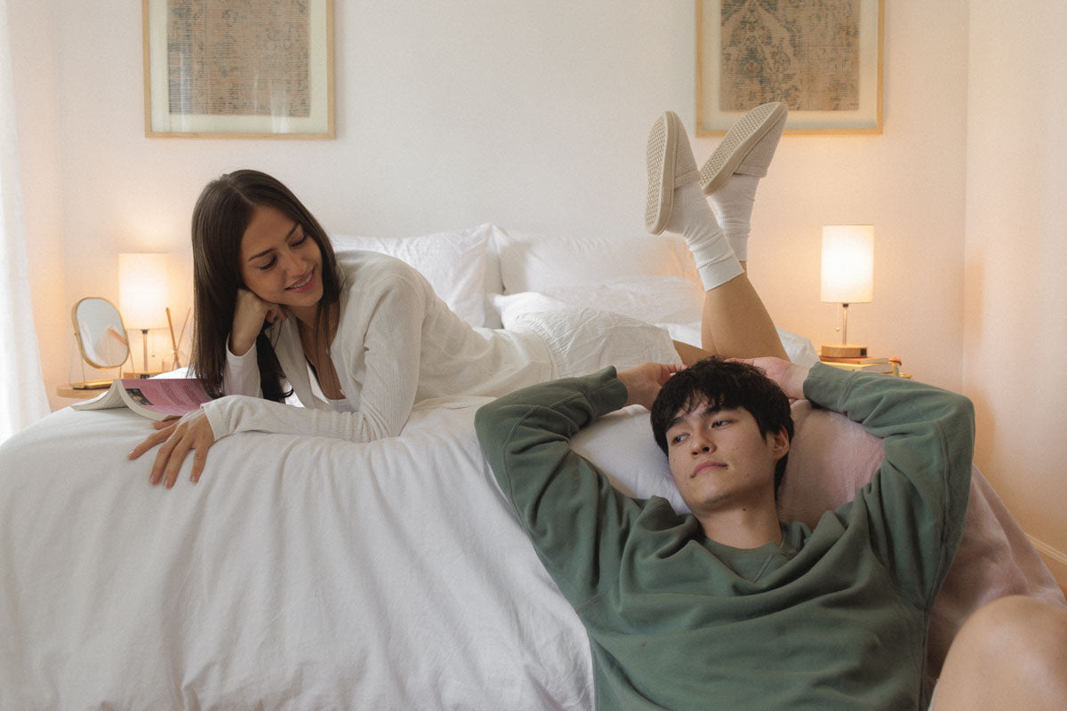 Woman reading on a white bed while a man lounges on the floor beside her in a cozy bedroom with warm lamps and framed art