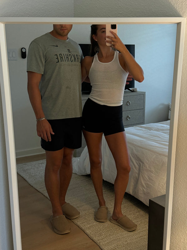 Two people taking a bedroom mirror selfie wearing dark brown corduroy slippers, woman in white tank and black shorts