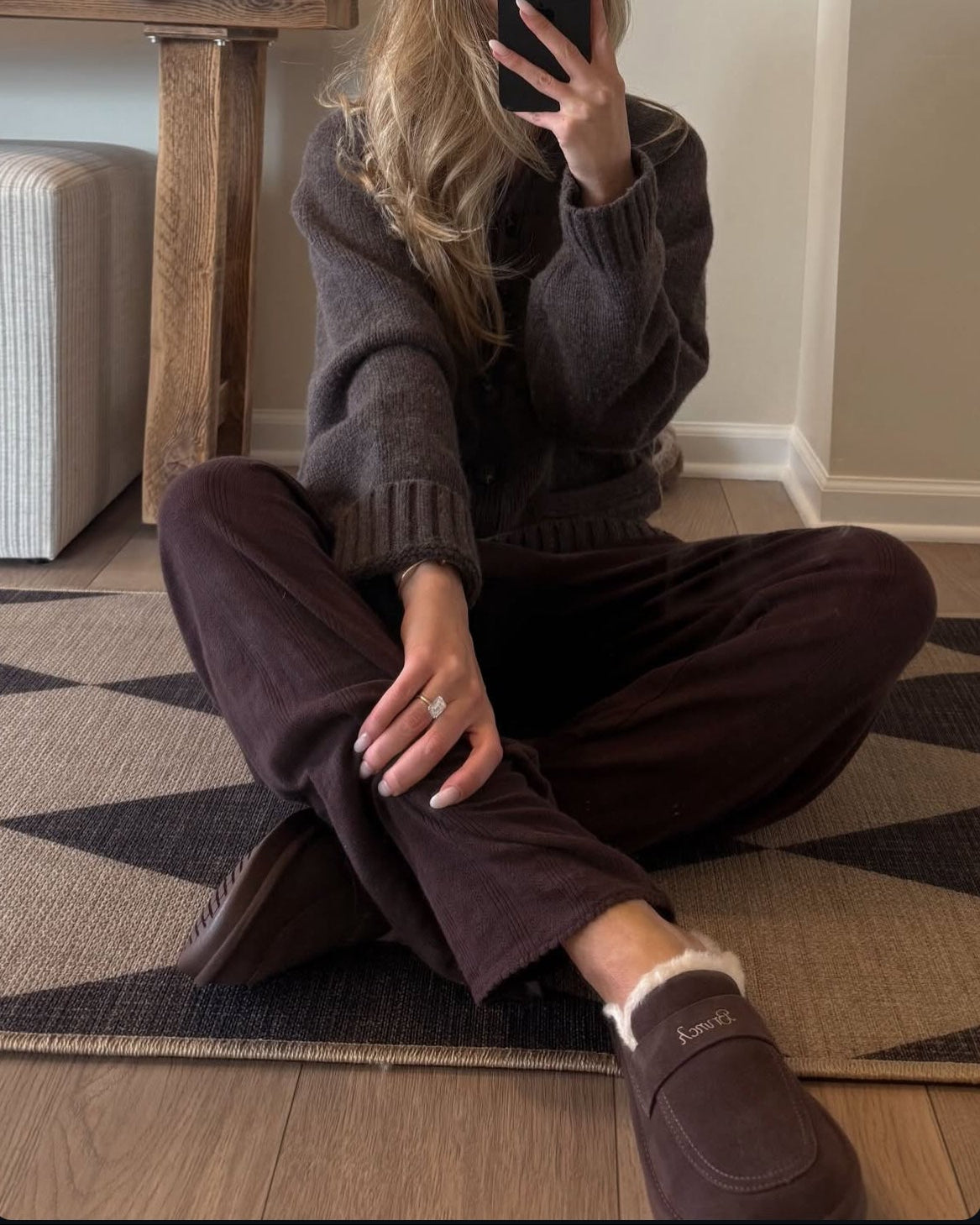 Woman taking a mirror selfie wearing a brown sweater and pants in a room with a wooden table and handbag.