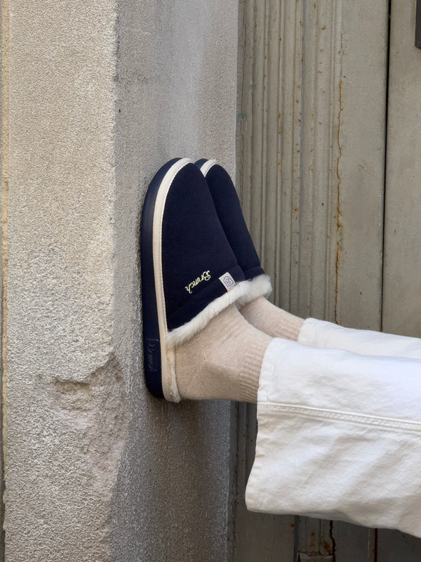 Navy slip-on slippers with white faux fur trim and beige socks, feet resting against a textured wall; embroidered text: "Brunch".