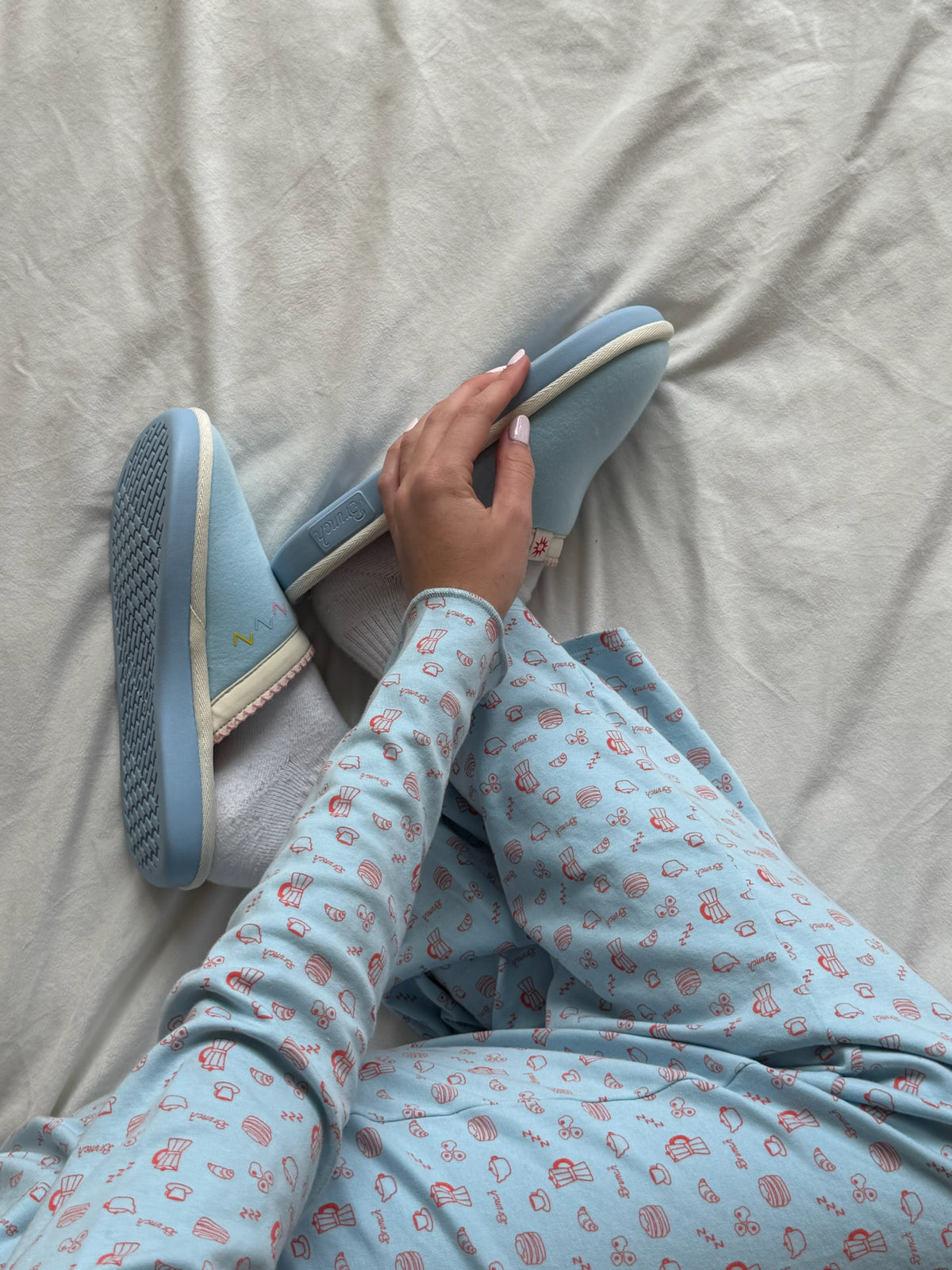 Person seated on pale sheet holding light-blue slip-on slippers, wearing light-blue pajama pants with small breakfast icons.