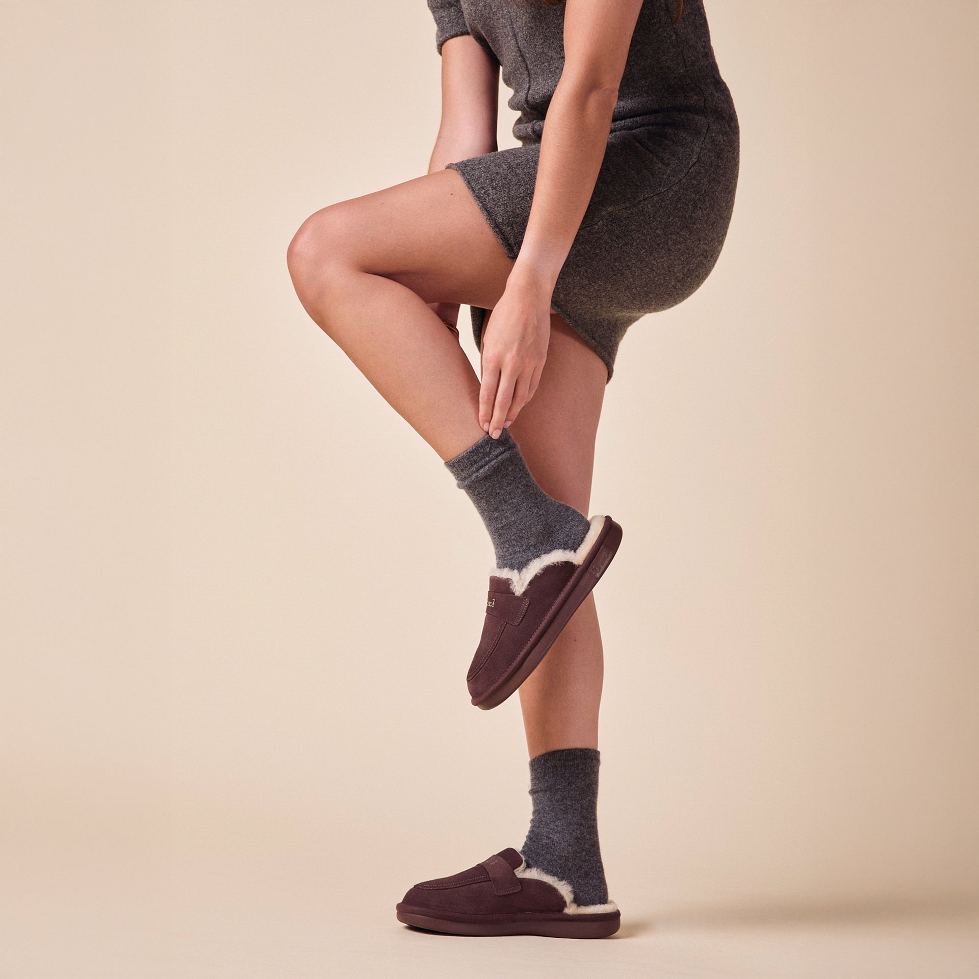 Woman wearing espresso-brown shearling-lined slippers with gray socks and a gray knit dress, one foot lifted