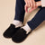 Black suede, shearling-lined slip-on slippers on feet wearing white socks and navy pants.