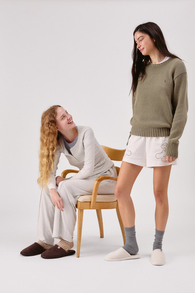 Two women modeling brown and cream terry slippers, one seated on a wooden chair smiling at a standing friend