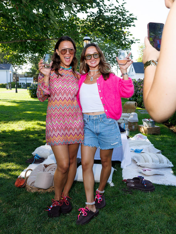 Two women raising wine glasses at a sunny backyard picnic, wearing dark brown slippers with pink laces