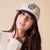 Young woman wearing cream baseball cap with navy brim and embroidered B logo and cream sweater