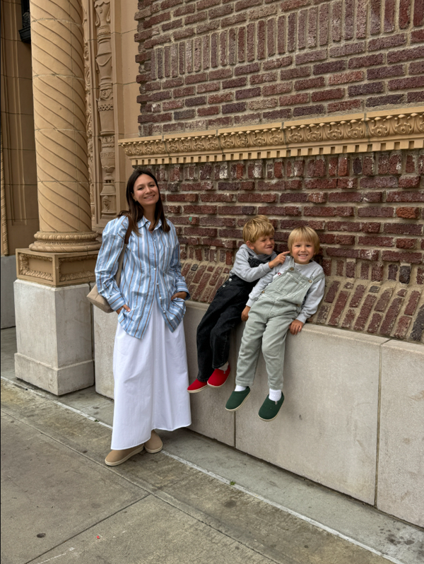 Woman standing beside two smiling young boys seated on a stone ledge against a decorative brick building