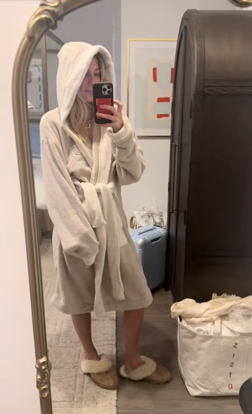 Person in a hooded beige bathrobe and slippers taking a mirror selfie in a bedroom; suitcase and laundry bag by an armoire.