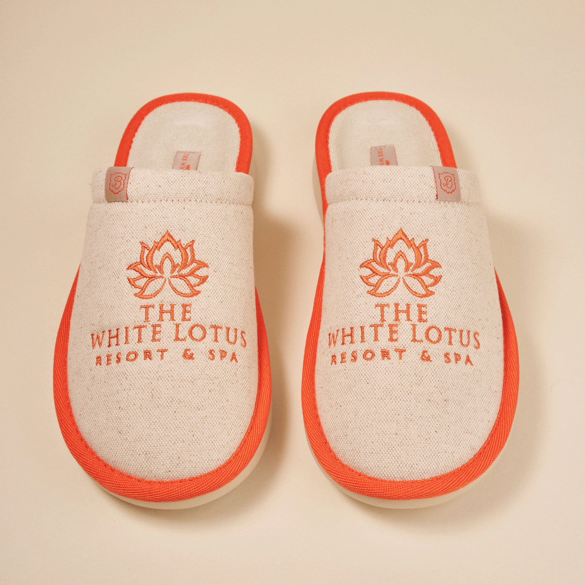 Pair of beige spa slippers with orange trim embroidered 'THE WHITE LOTUS RESORT & SPA' and lotus logo