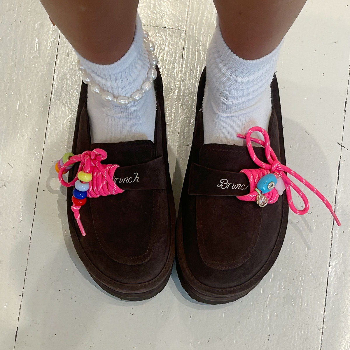 Top view of Carrie Charm Kit hot-pink paracord laces with colorful beads and charms threaded onto dark brown suede loafers on white floor
