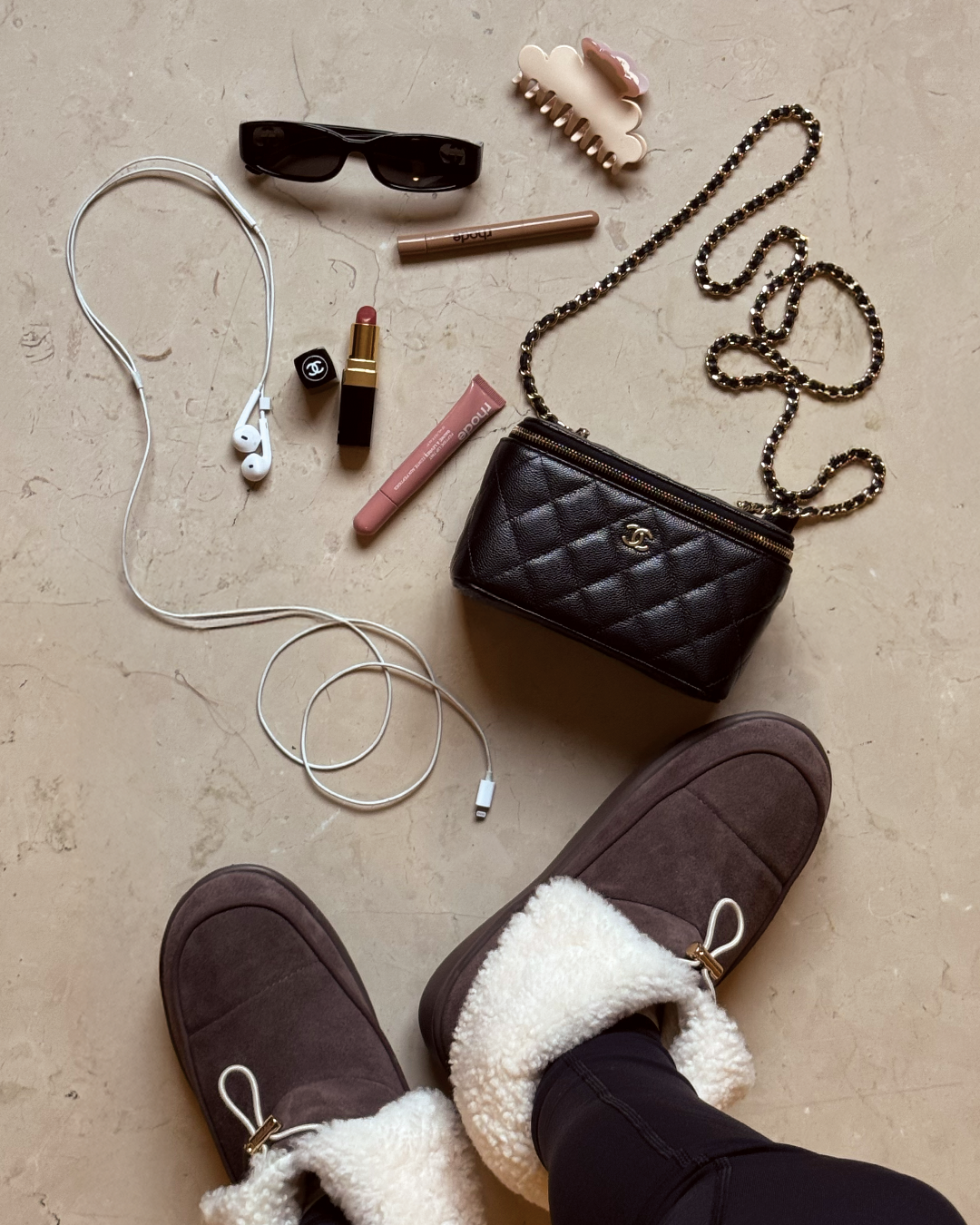 Flat lay: quilted black chain purse, sunglasses, earbuds with cable, lipstick and gloss, hair clip, shearling slippers.