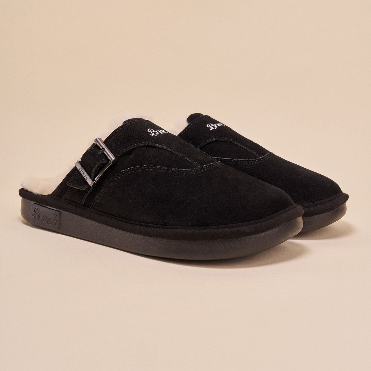 Front three-quarter view of Amour Shearling slip-on mule in black suede showcasing shearling lining, adjustable side buckle, and Brunch-branded sole on beige background