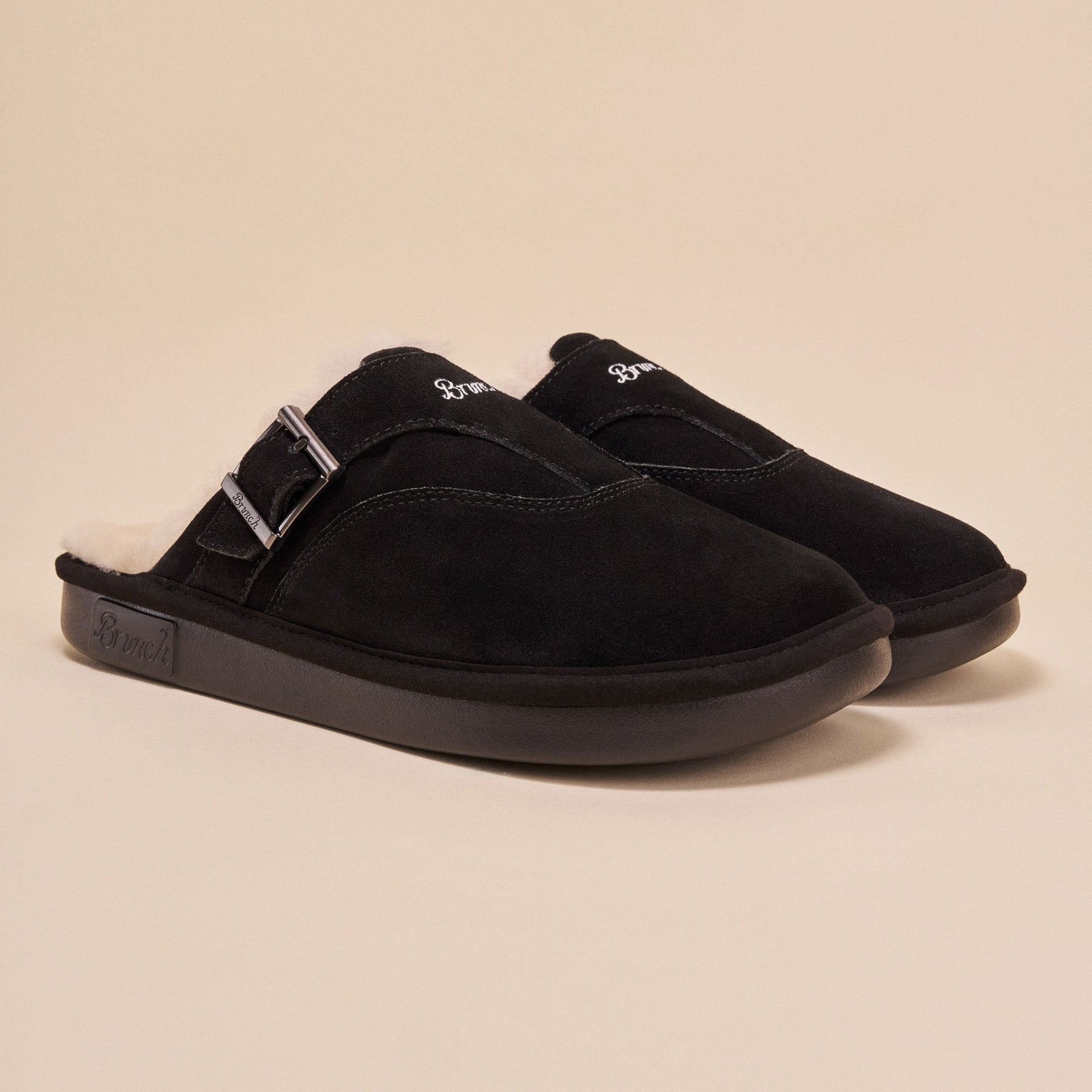 Front three-quarter view of Amour Shearling slip-on mule in black suede showcasing shearling lining, adjustable side buckle, and Brunch-branded sole on beige background