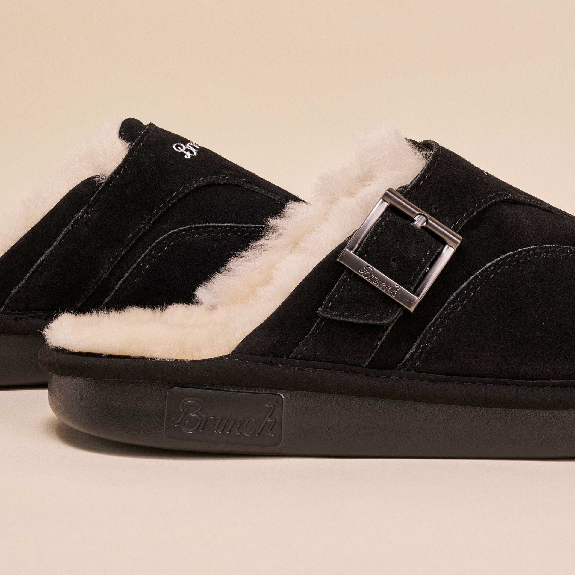 Side view of Amour Shearling slipper in black suede with cream lining, adjustable buckle, and Brunch logo on sole