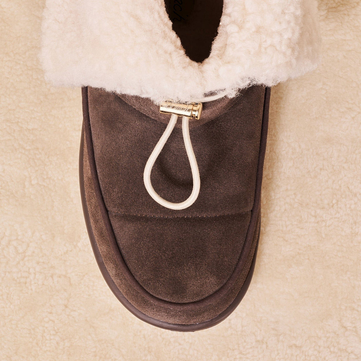 Top view of Apres Boot in dark brown suede showing folded shearling collar and front toggle loop on cream sherpa background