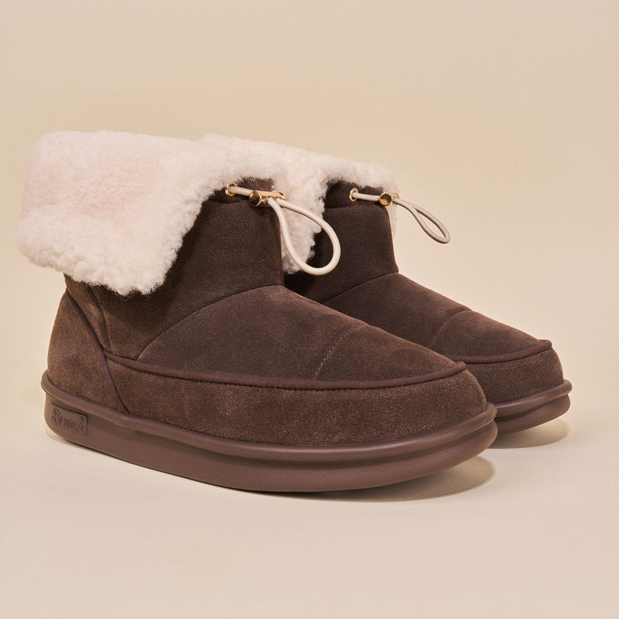Three-quarter view of Apres Boot dark brown suede ankle boots with cream shearling cuff and toggle drawcord on beige background