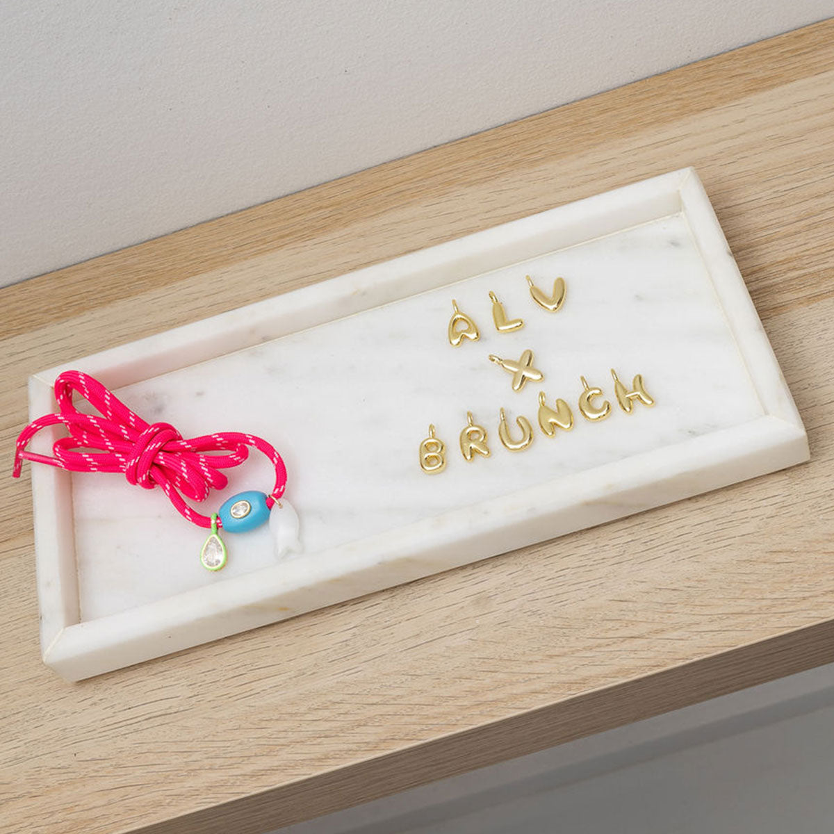Top-down view of Carrie charm kit showcasing gold letter charms spelling ALV x BRUNCH and a neon pink cord with enamel and teardrop charms on a white marble tray