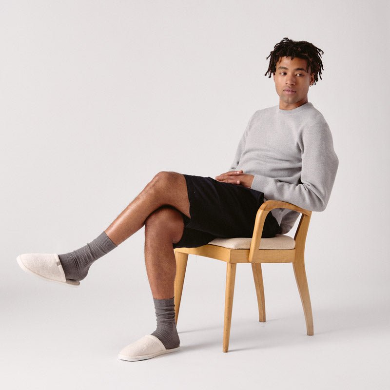 Model seated in a wooden chair wearing cream terry Classic slippers with a slim profile and gray socks on a light studio background