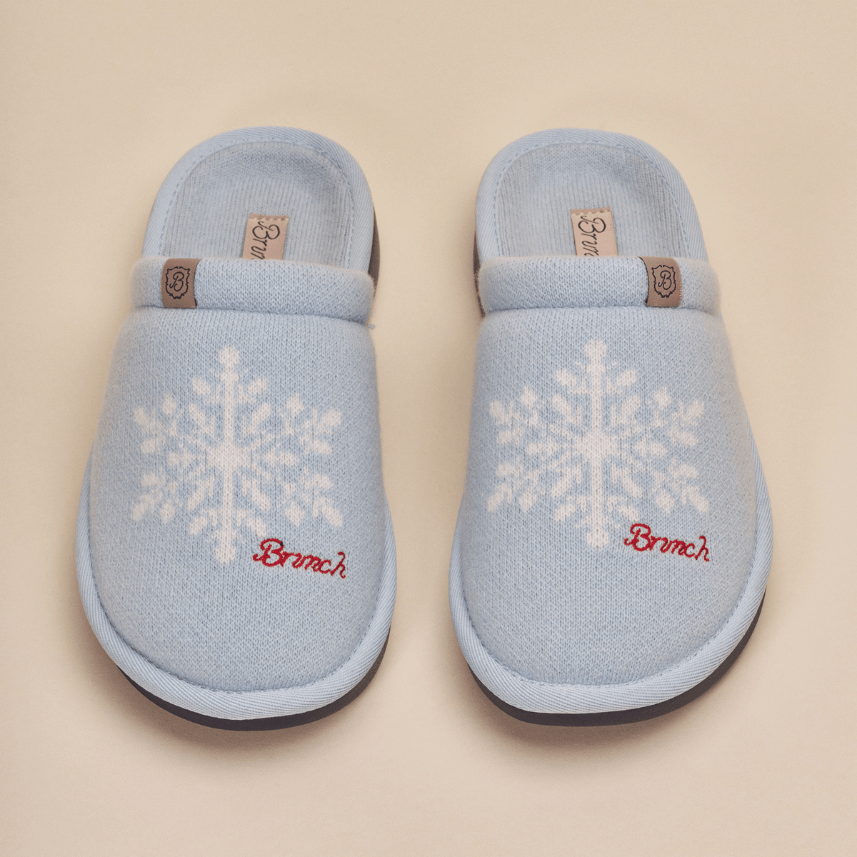 Top view of Essential Cashmere slippers in blue showing white snowflake knit pattern, red Brunch embroidery and leather logo tab on a beige background