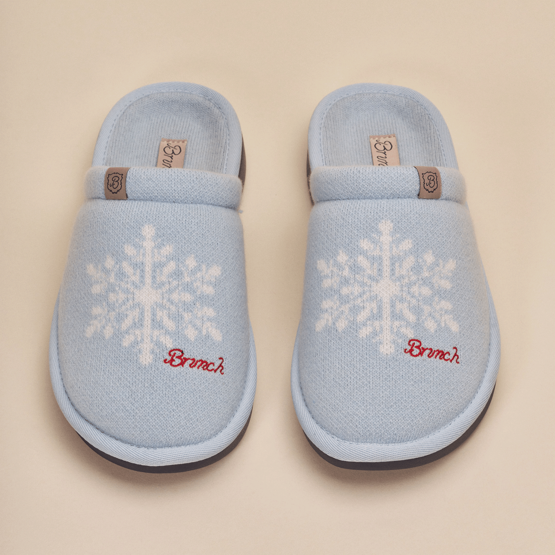 Top view of Essential Cashmere slippers in blue showing white snowflake knit pattern, red Brunch embroidery and leather logo tab on a beige background
