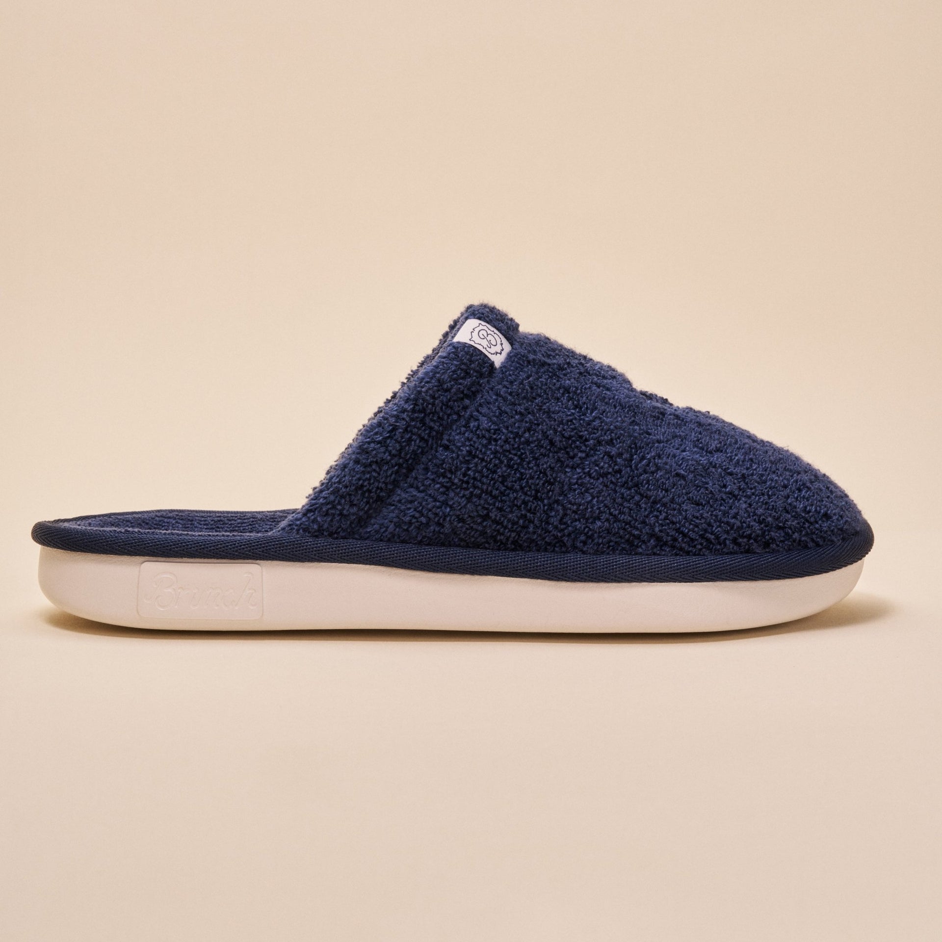 Side view of navy terry open-back slipper showcasing plush terry upper and white cushioned sole on beige background