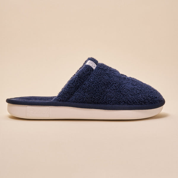 Side view of navy terry open-back slipper showcasing plush terry upper and white cushioned sole on beige background