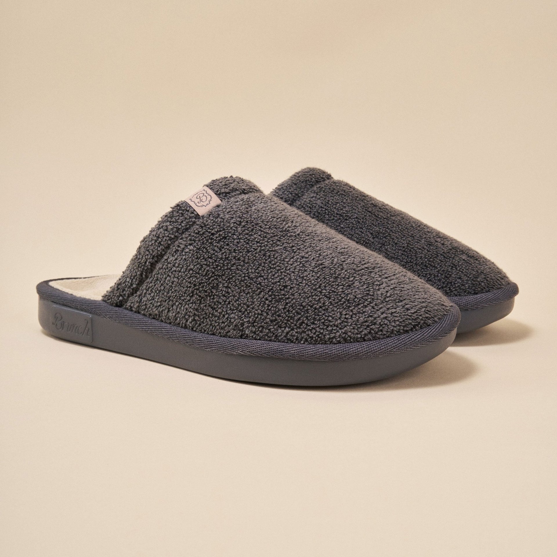 Angled view of Essential slipper, dark grey terry, closed-toe padded upper and molded EVA sole on beige background