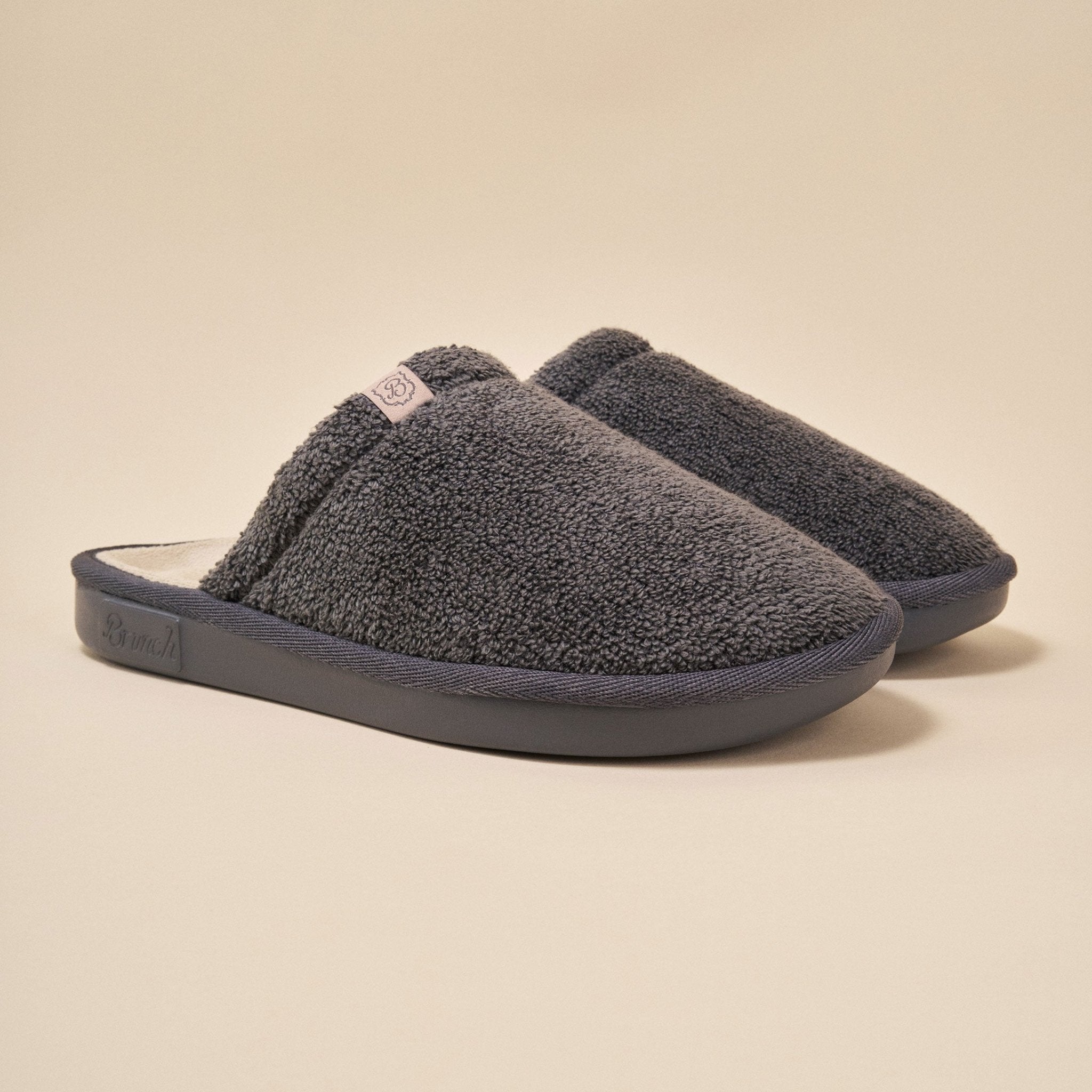Angled view of Essential slipper, dark grey terry, closed-toe padded upper and molded EVA sole on beige background