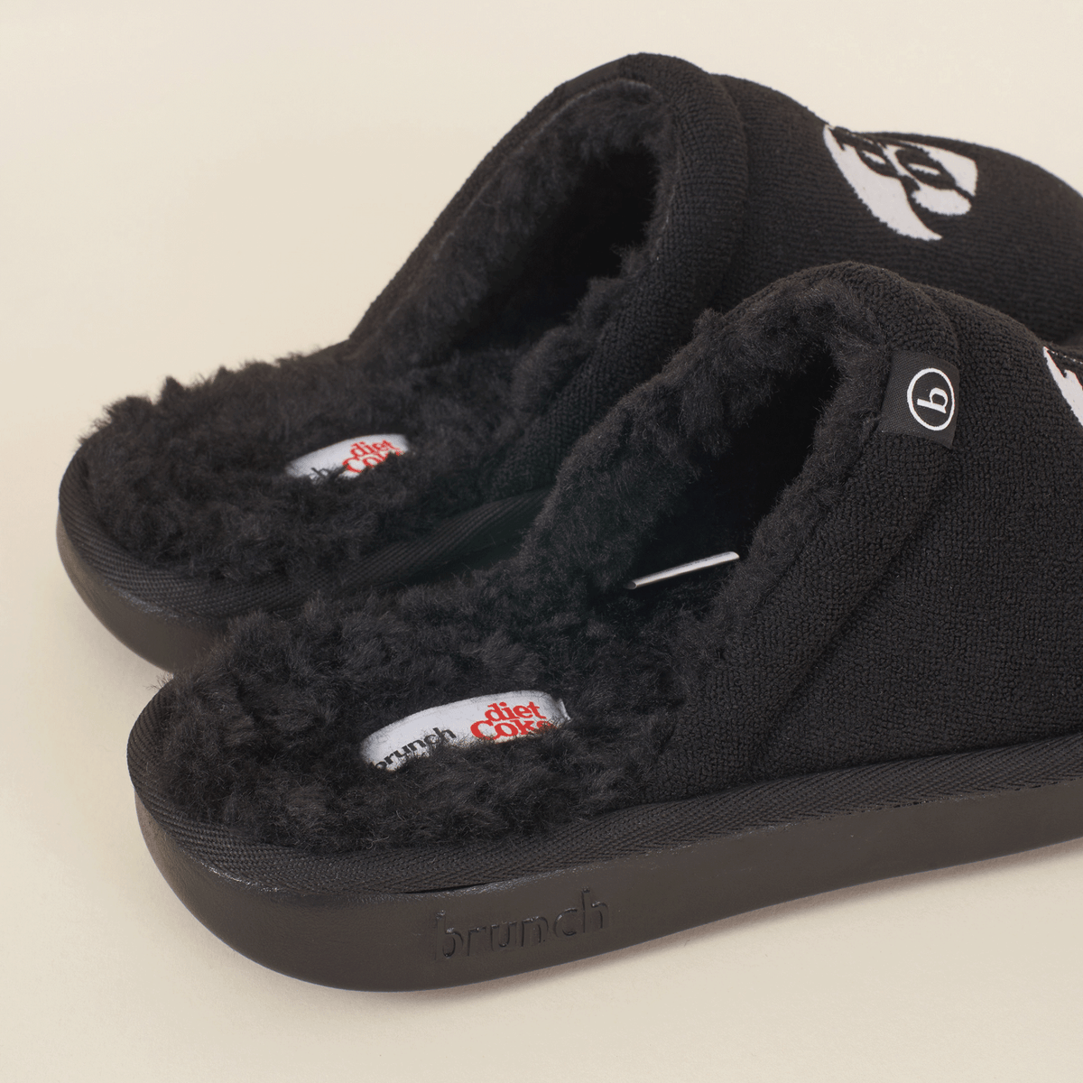 Three-quarter view of black slip-on slippers with faux shearling lining, Diet Coke footbed label and embossed Brunch sole