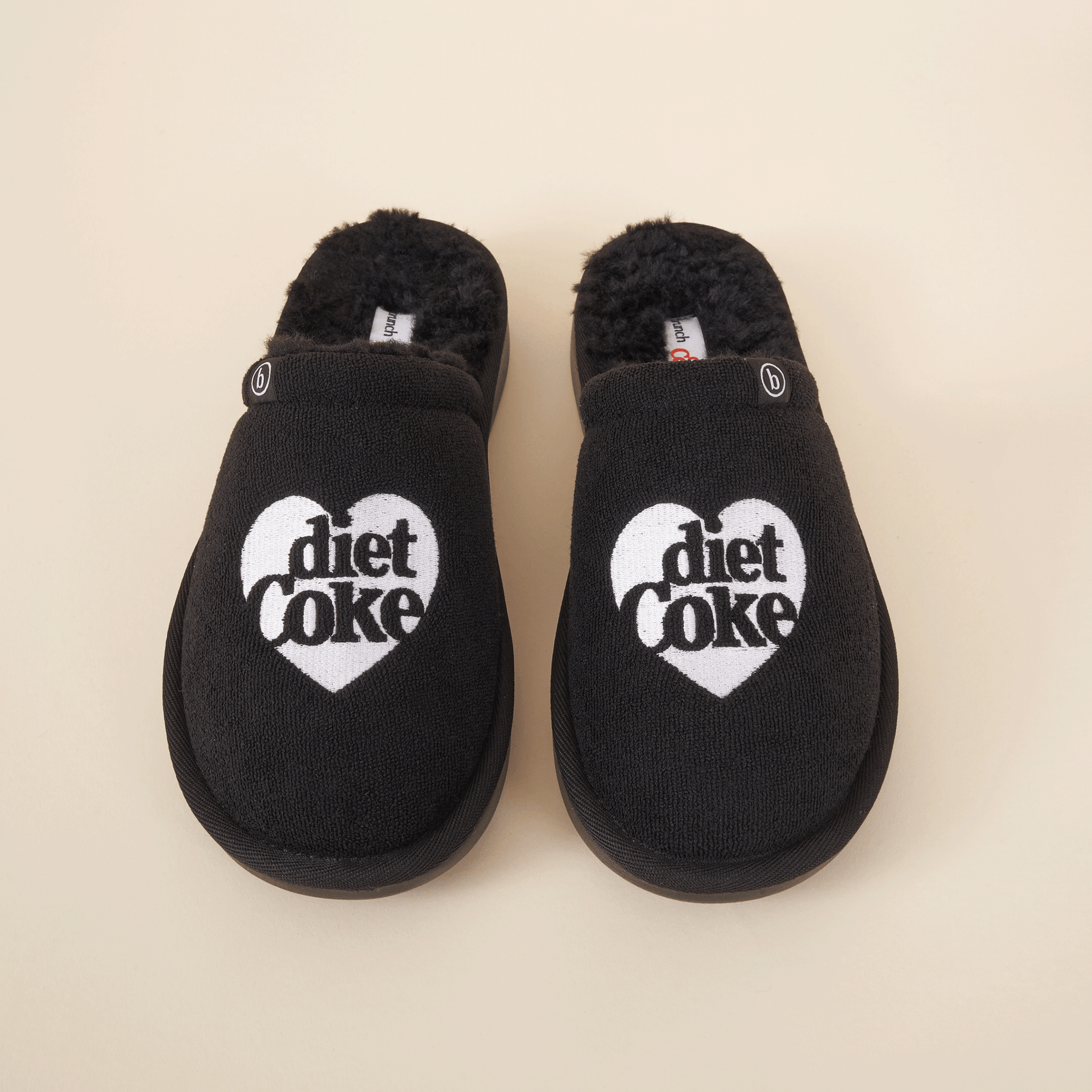 Top-down view of black shearling slippers showcasing white heart-shaped Diet Coke embroidery and faux-shearling lining on cream background