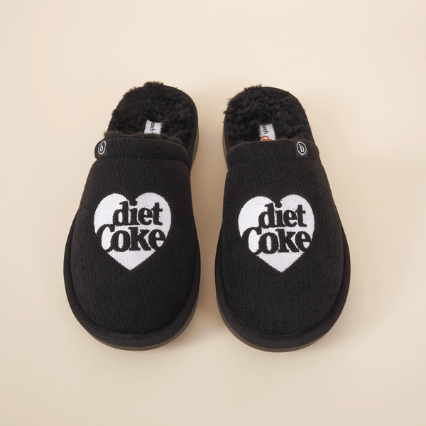 Top-down view of black shearling slippers showcasing white heart-shaped Diet Coke embroidery and faux-shearling lining on cream background