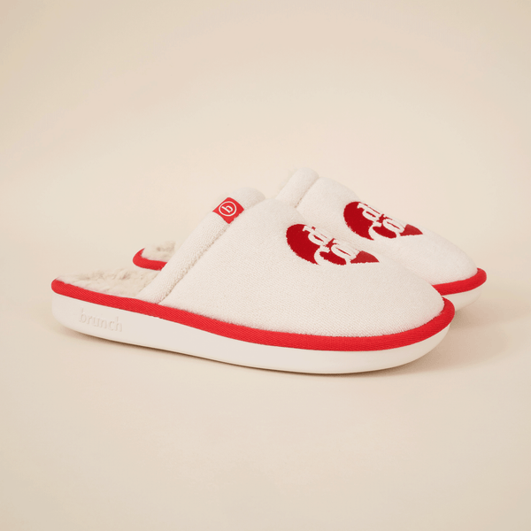 Three-quarter view of cream slip-on slippers showing red Diet Coke logo, red piping and faux-shearling lining on beige
