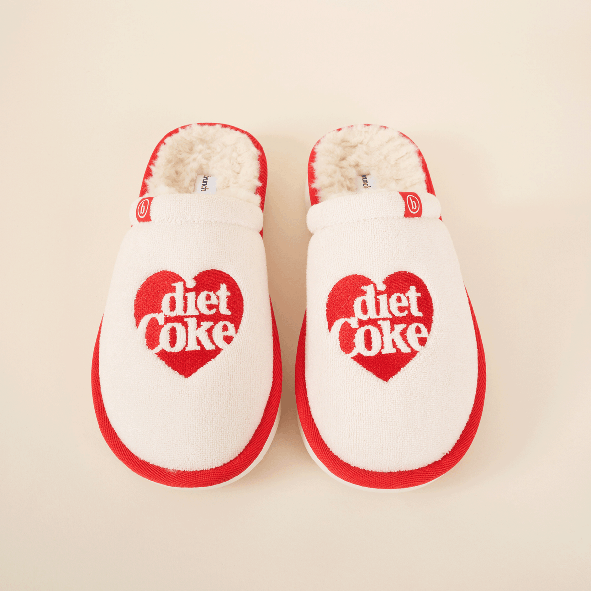 Top-down view of cream and red Diet Coke shearling slip-on slippers showcasing red heart "diet coke" embroidery and faux shearling lining on cream background