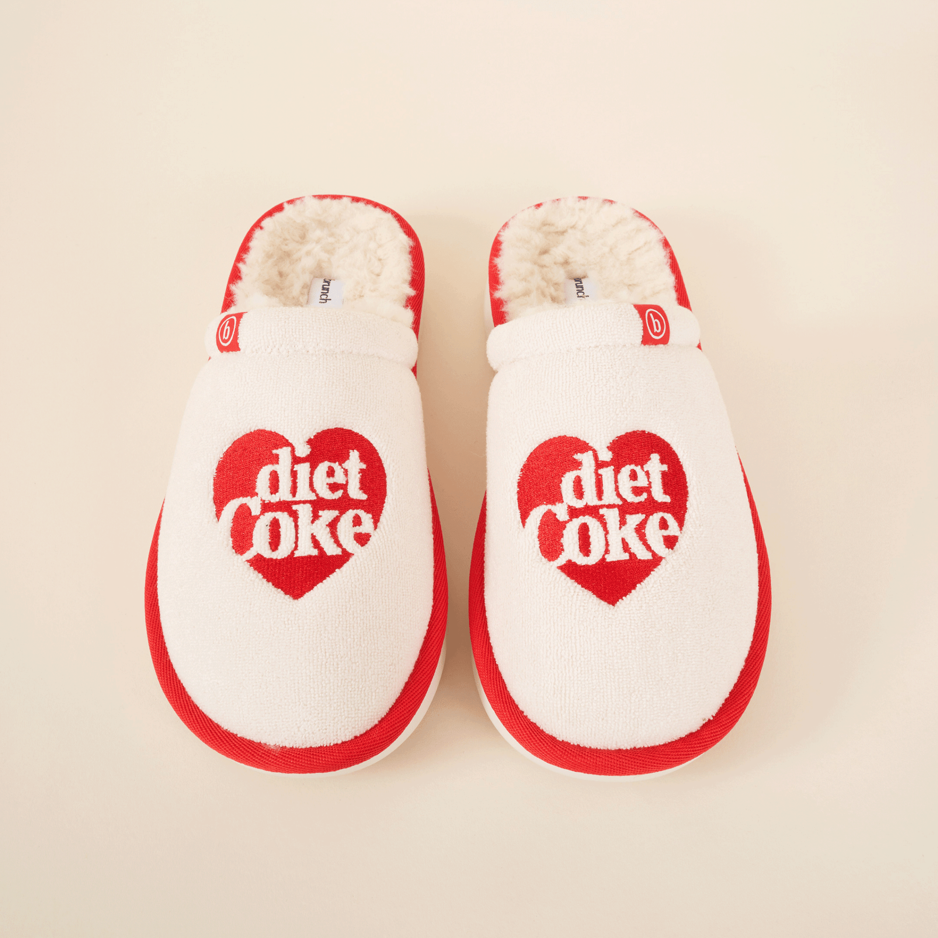 Top-down view of cream and red Diet Coke shearling slip-on slippers showcasing red heart "diet coke" embroidery and faux shearling lining on cream background