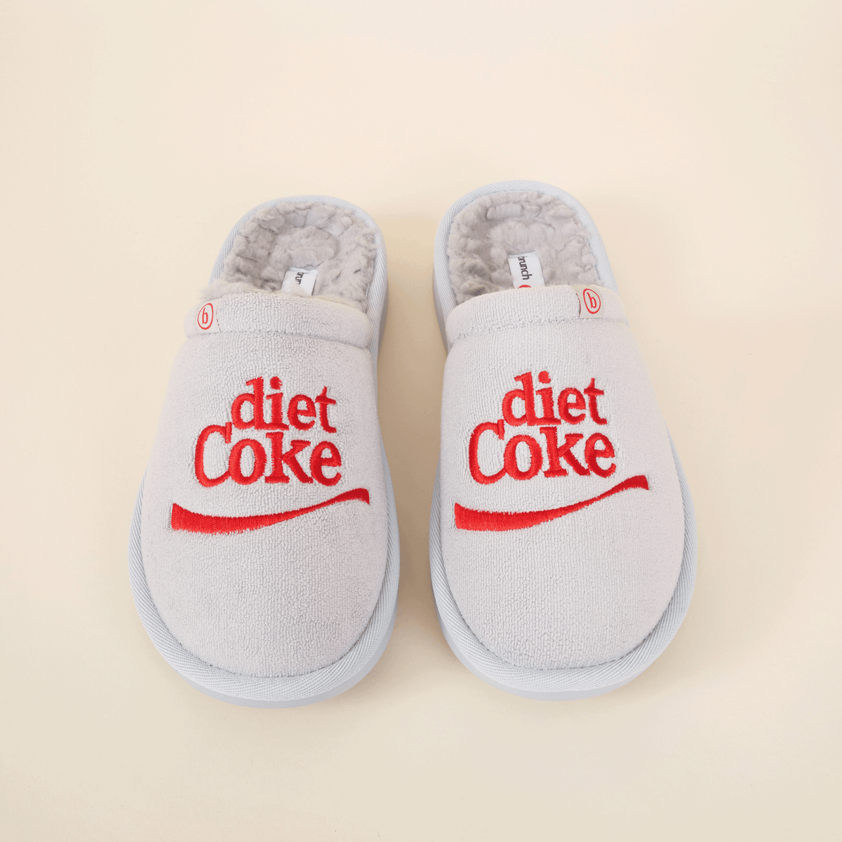 Top-down view of grey shearling slippers with red embroidered diet Coke logo on beige background