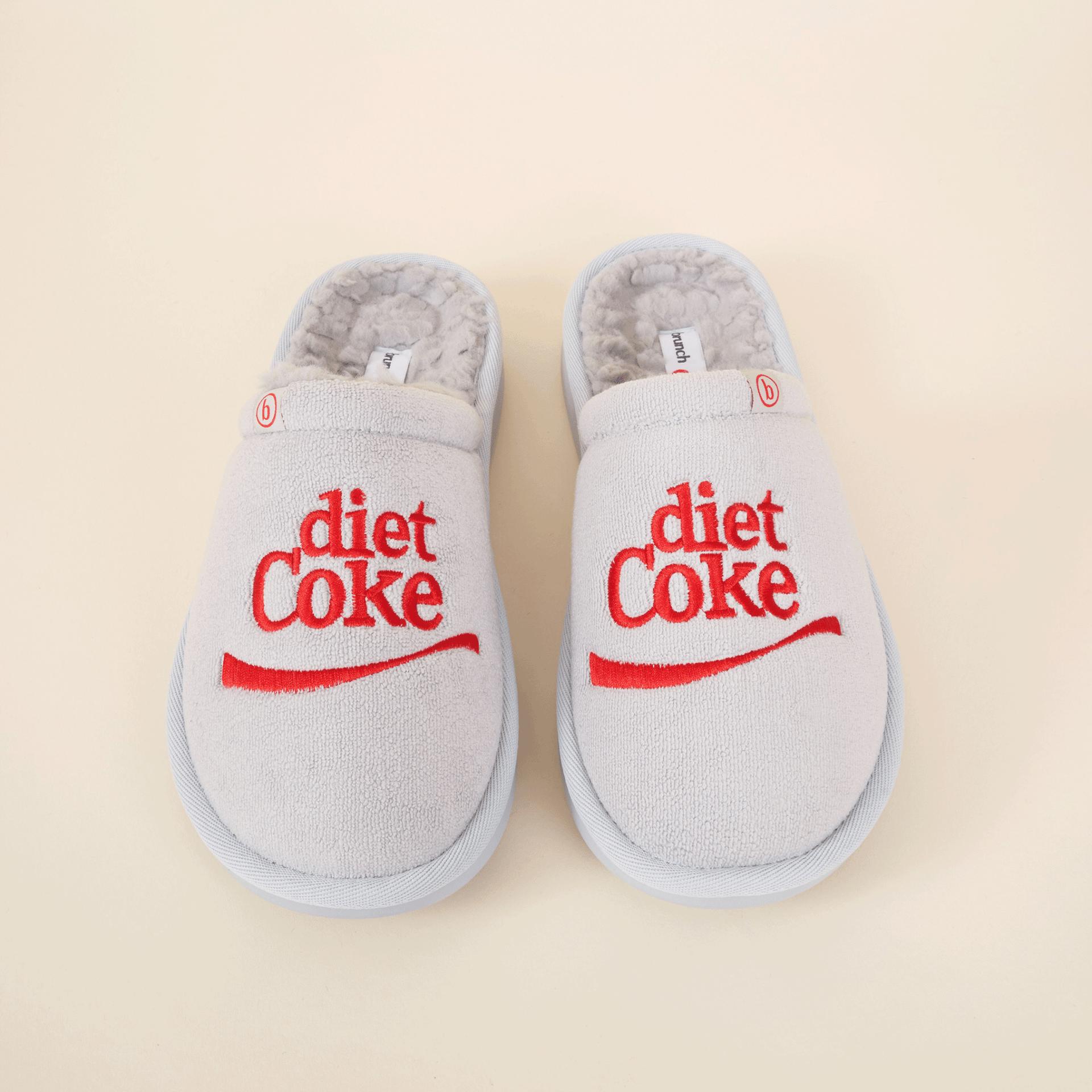 Top-down view of grey shearling slippers with red embroidered diet Coke logo on beige background