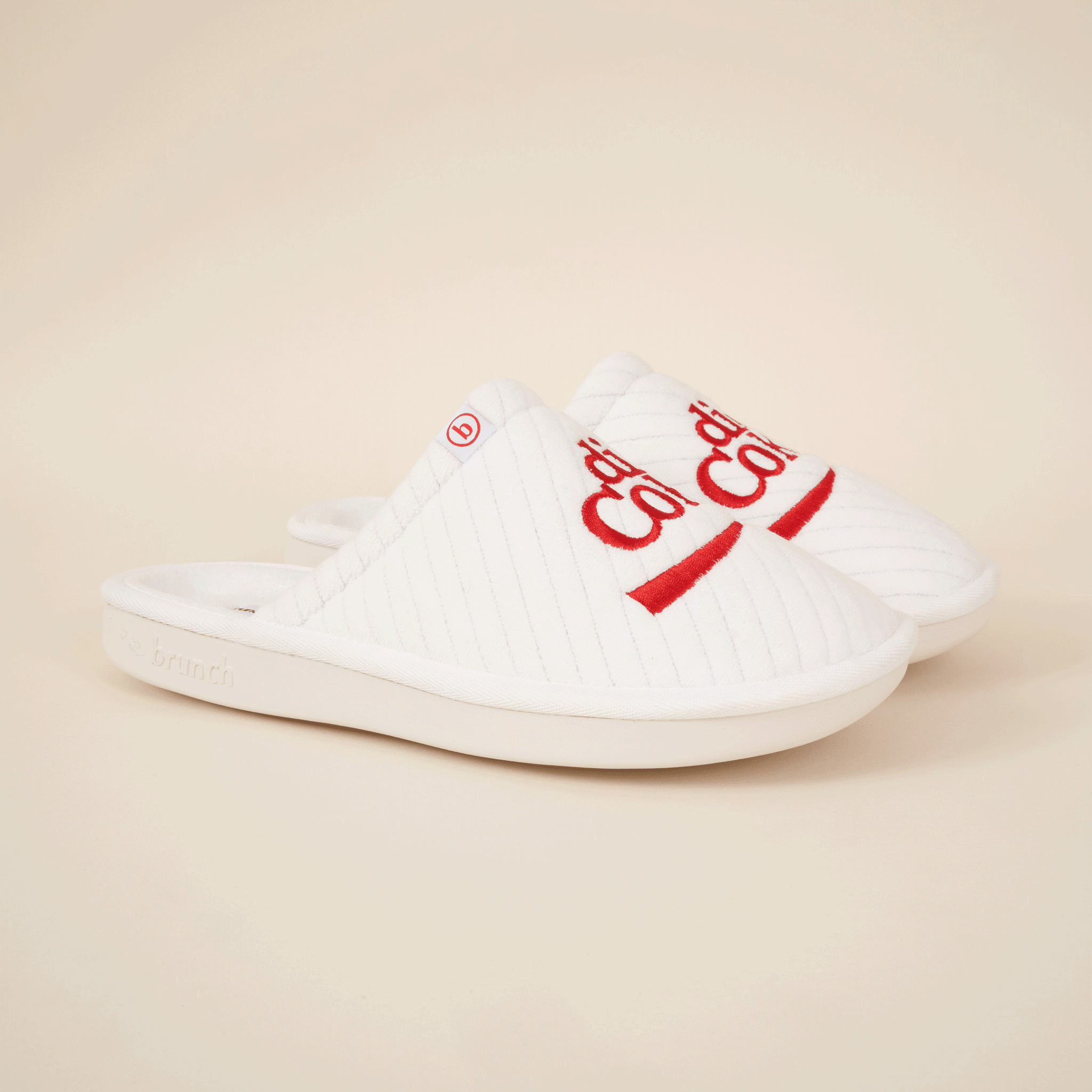 Angled view of white Brunch x Diet Coke slippers with red embroidered Diet Coke logo and quilted upper on beige background