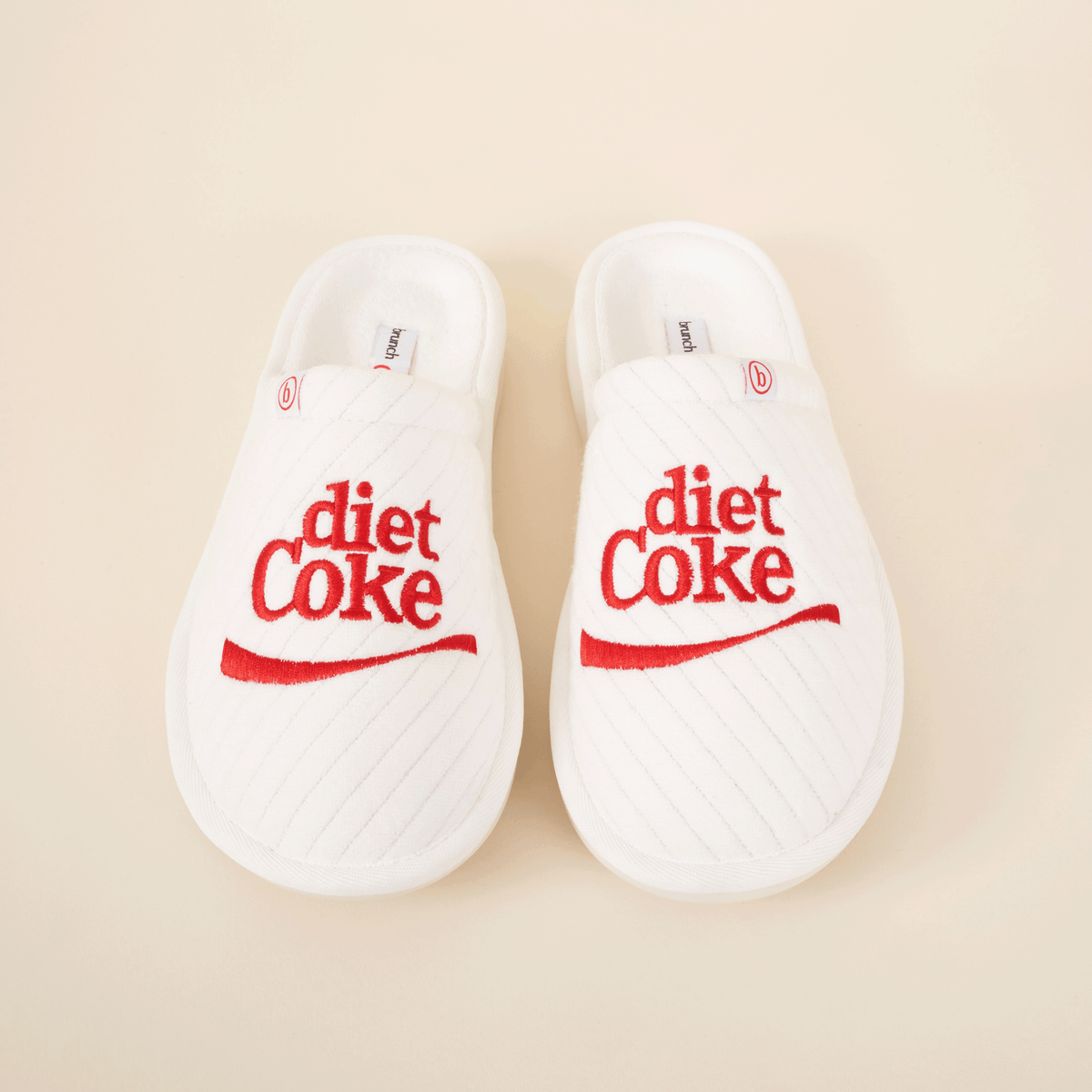 Overhead view of white L'Essentiel slippers showcasing red embroidered diet Coke logo and quilted upper on beige background
