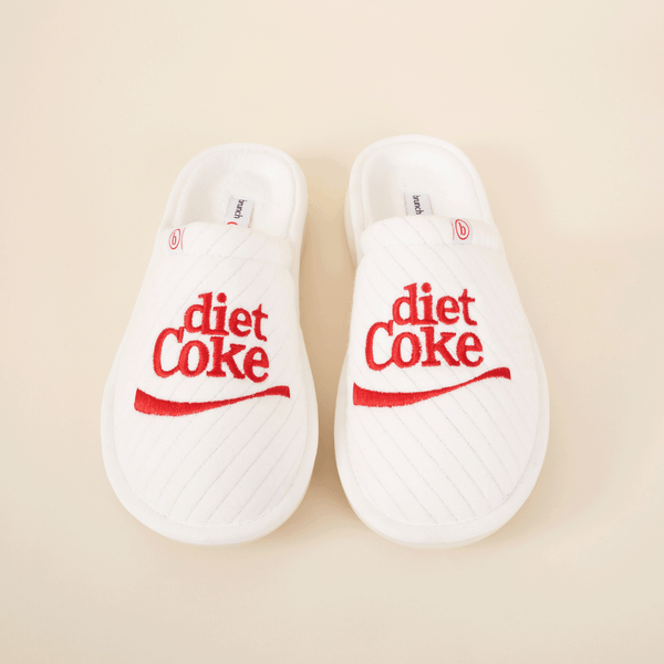 Overhead view of white L'Essentiel slippers showcasing red embroidered diet Coke logo and quilted upper on beige background