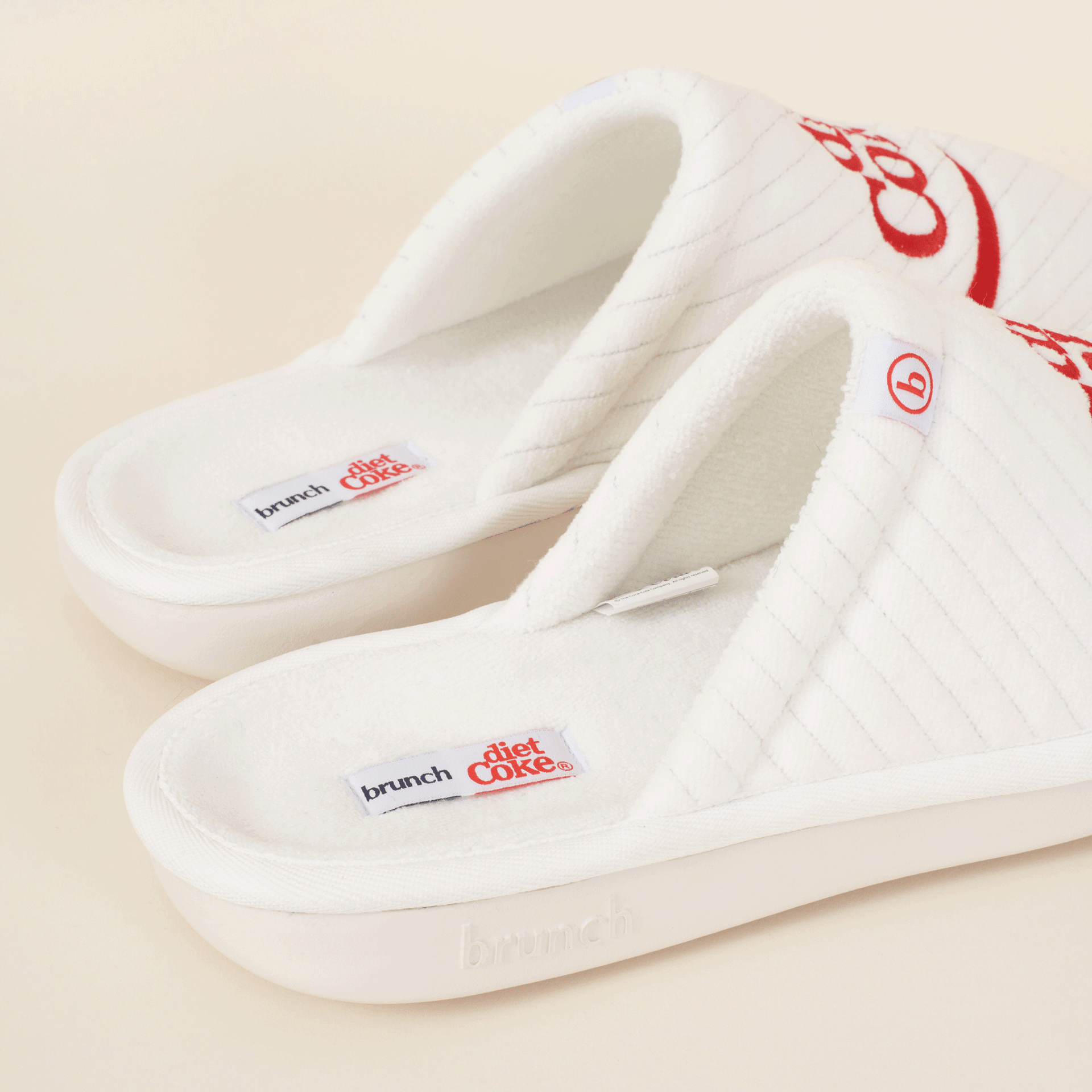 Three-quarter view of white Diet Coke x Brunch quilted slippers with red embroidered logo, terry footbed labels, and cushioned platform on a beige background