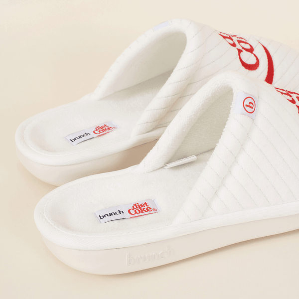 Three-quarter view of white Diet Coke x Brunch quilted slippers with red embroidered logo, terry footbed labels, and cushioned platform on a beige background