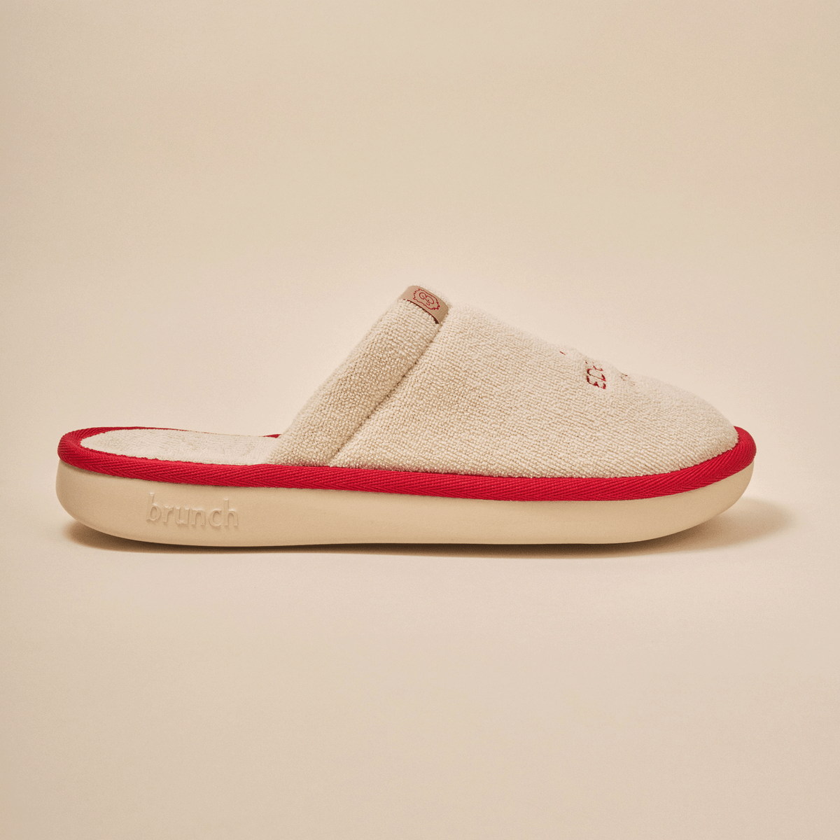 Side view of Essential Eden Rock slipper in beige with soft terry upper, red trim and Eden Roc embroidery on neutral backdrop