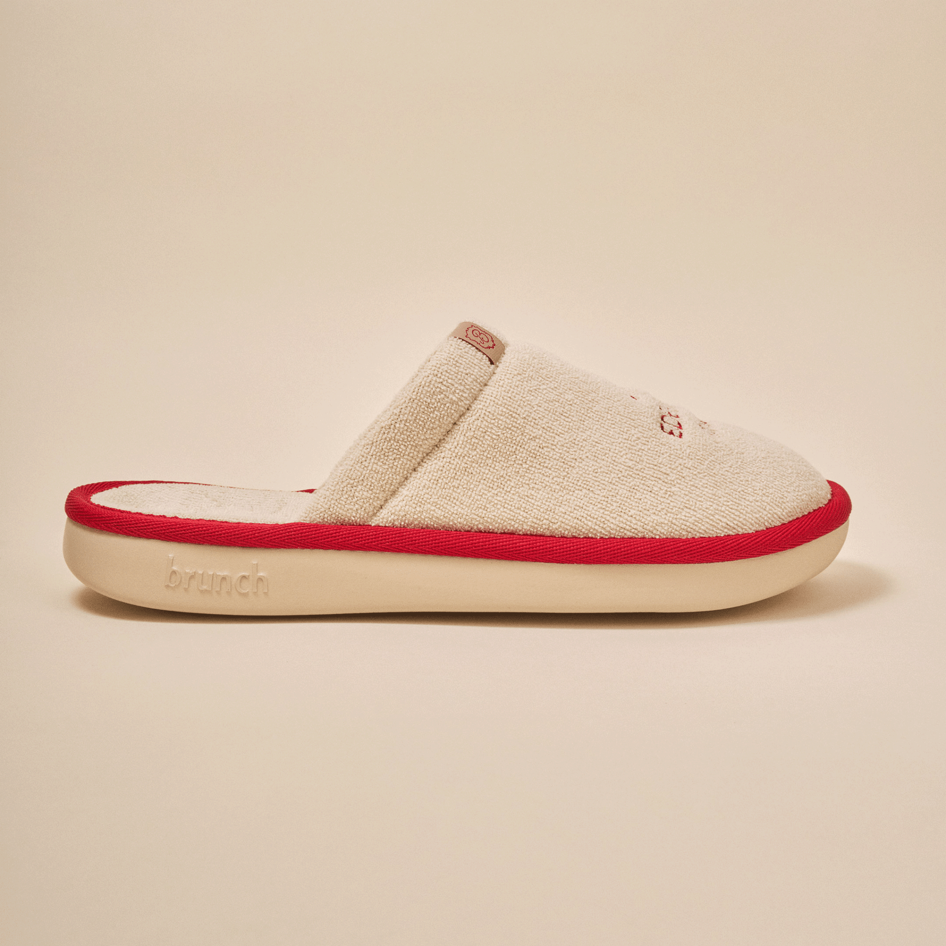 Side view of Essential Eden Rock slipper in beige with soft terry upper, red trim and Eden Roc embroidery on neutral backdrop