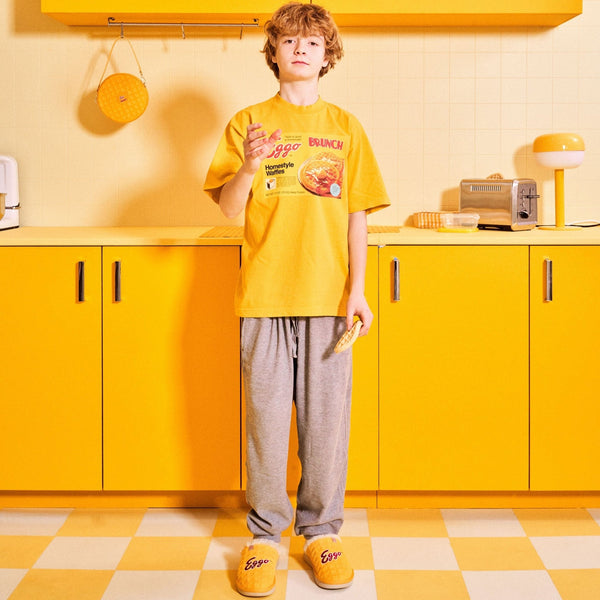 Person standing in a yellow kitchen wearing yellow Eggo slippers and a matching Eggo t-shirt while holding a waffle