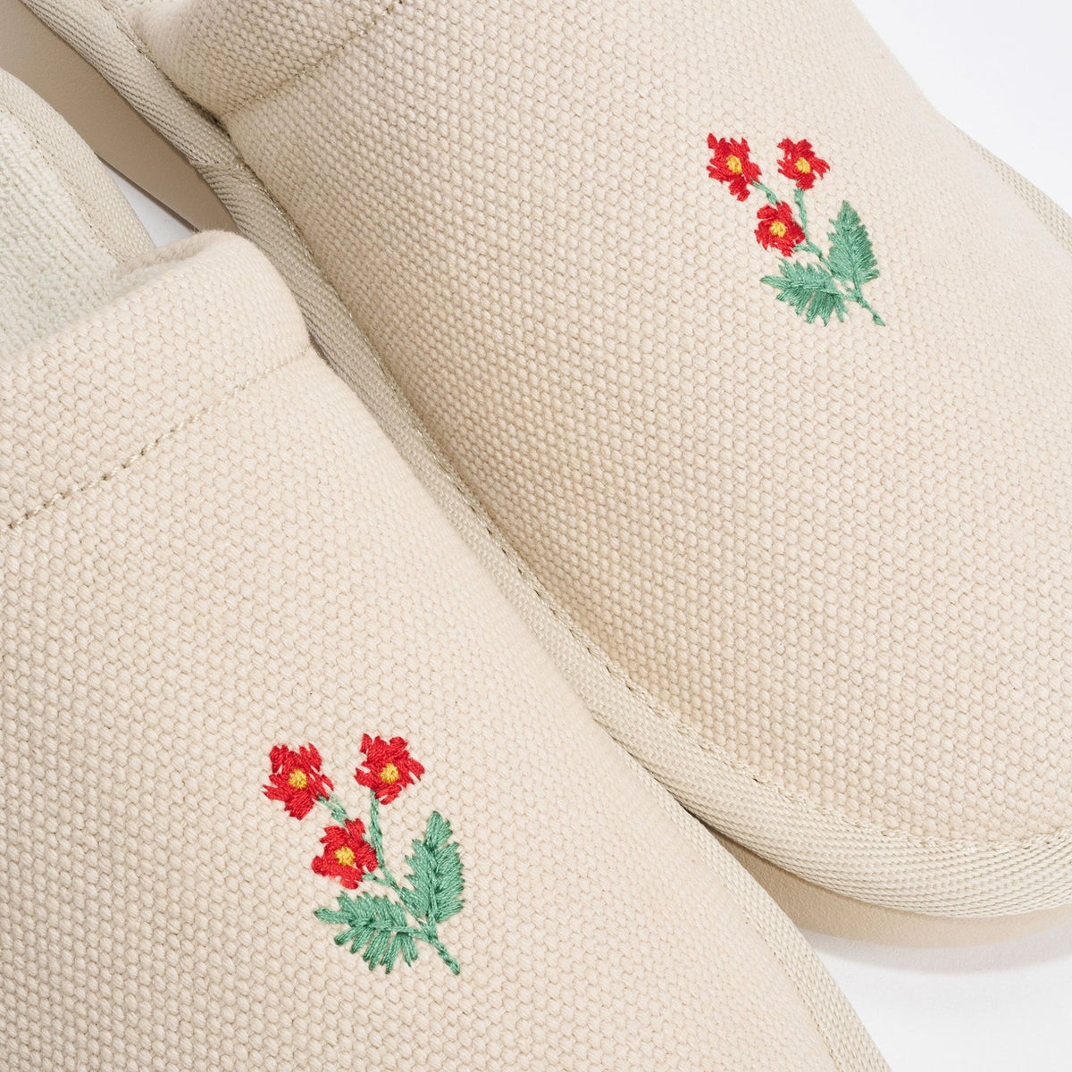 Top view of pair of beige canvas slippers showcasing hand stitched red wildflower embroidery on toes with white background