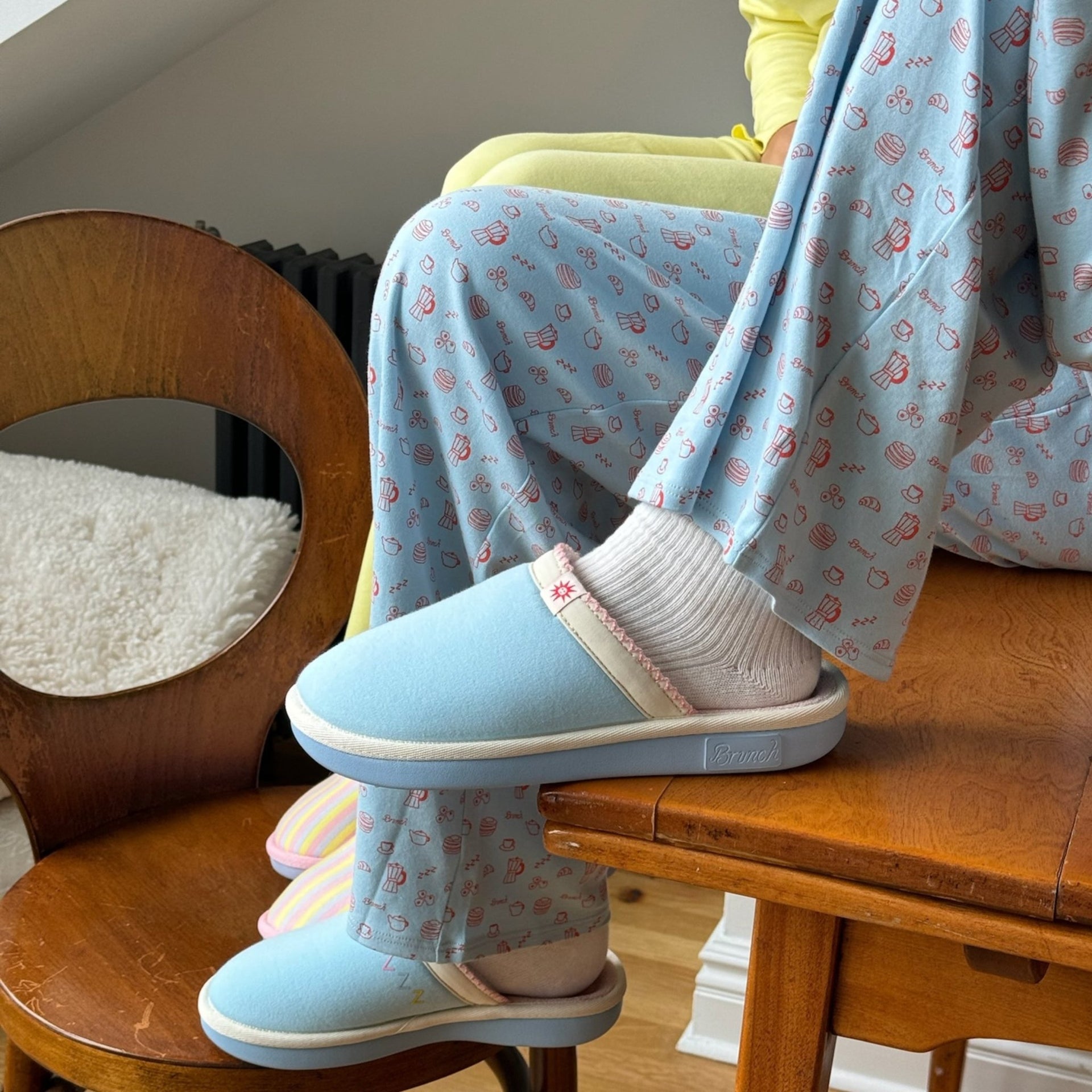 Side view of Essential Cozyland slipper in French Blue showing cushioned sole and trim, worn with patterned pajamas on a wooden chair