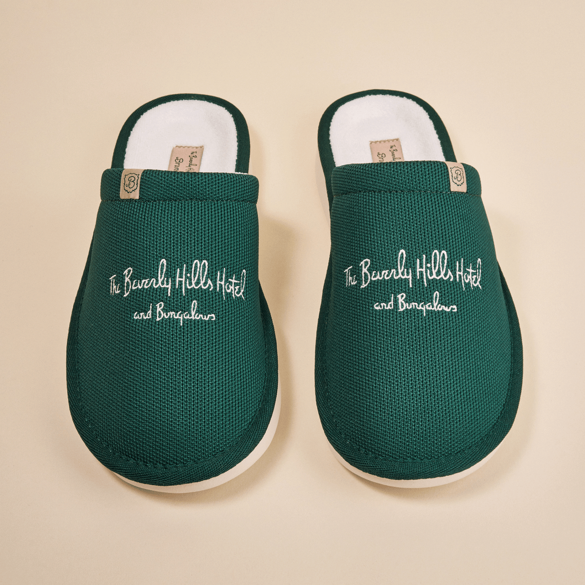 Top-down view of green Essential slippers embroidered with The Beverly Hills Hotel and Bungalows text on a beige background