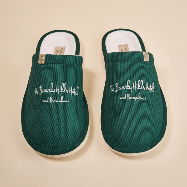 Top-down view of green Essential slippers embroidered with The Beverly Hills Hotel and Bungalows text on a beige background