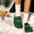 Front of green Essential Beverly Hills Hotel slippers with embroidered hotel logo on knit upper, worn with white socks on pink satin sheets